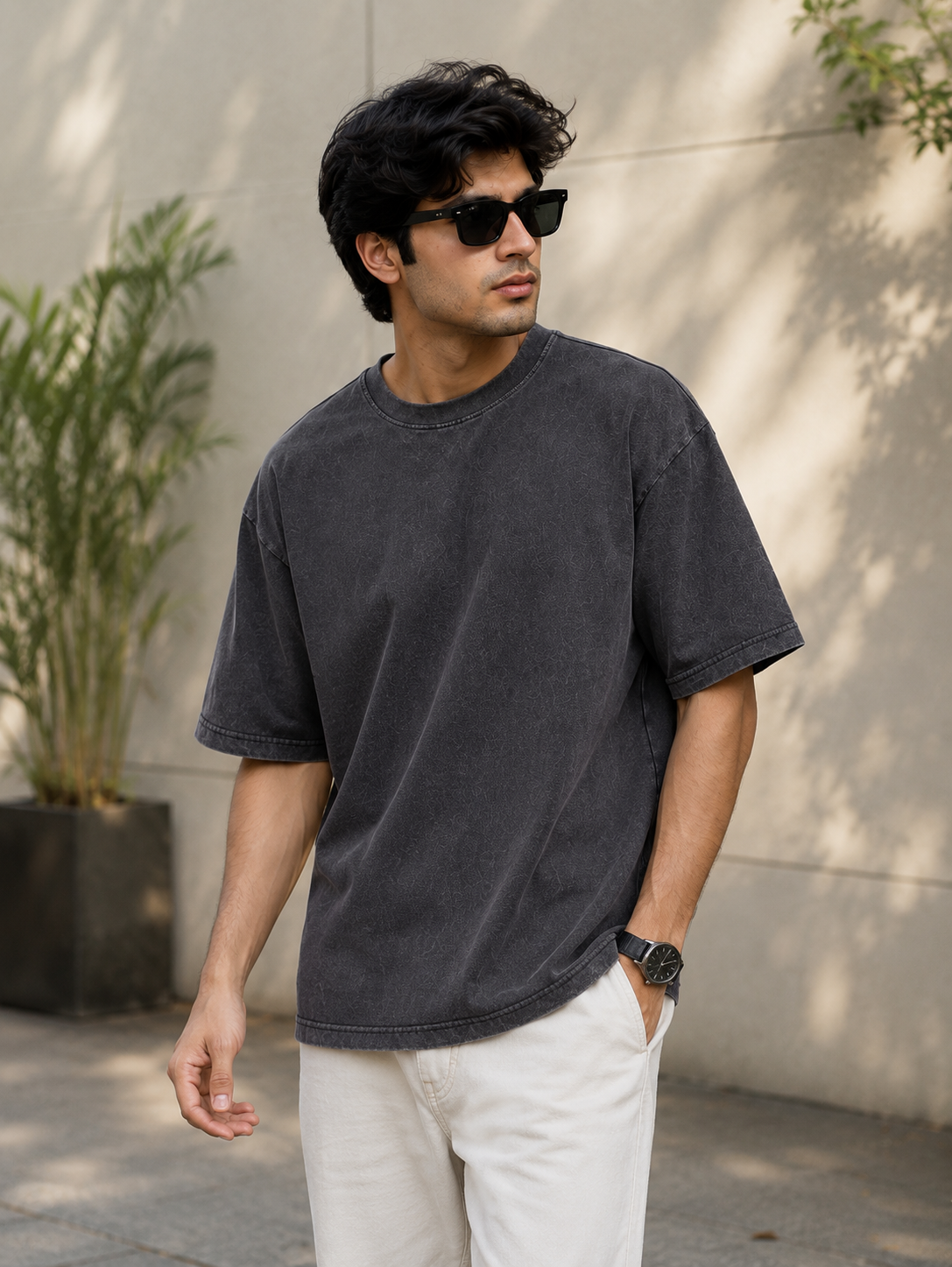 A man wearing a faded black washed cotton t-shirt and white pants, standing outdoors against a beige wall with shadows.