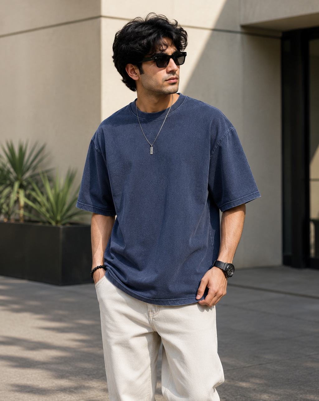 A man wearing an oversized navy blue t-shirt, off-white trousers, black sunglasses, and a silver necklace, standing outdoors in front of a modern beige building.
