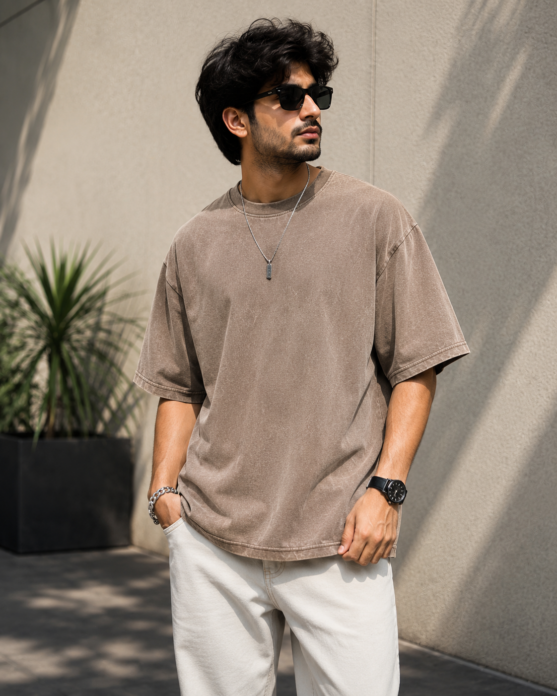 A man wearing an oversized, faded taupe-brown short-sleeve t-shirt, white trousers, sunglasses, and a necklace, standing against a textured concrete wall.