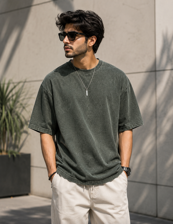 A man wearing a faded dark green short-sleeve t-shirt, cream-colored pants, and black sunglasses, standing outdoors against a light gray tiled wall.