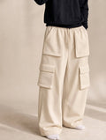 A person wearing loose, wide-leg cargo pants in a warm off-white or cream color, paired with a black long-sleeve top and white socks, standing against a neutral beige studio background.