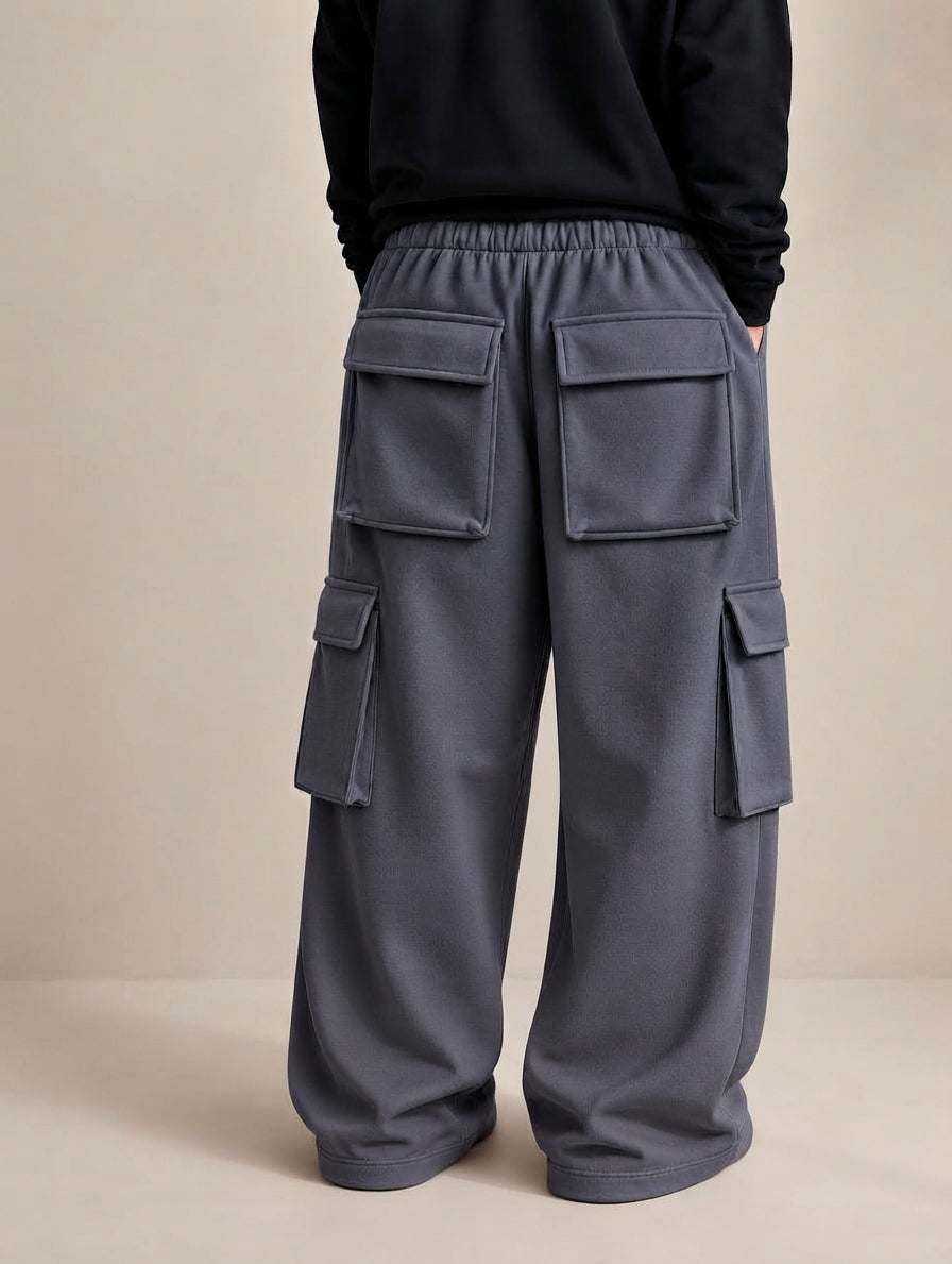 A person shown from the back wearing loose, wide-leg cargo pants in dark gray with an elastic waistband and multiple flap cargo pockets, paired with a black long-sleeve top against a plain beige studio background.