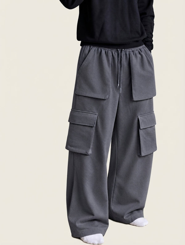 A person wearing loose-fit gray cargo pants with a drawstring waist, large flap cargo pockets, and a black long-sleeve top, shown against a plain light beige background.