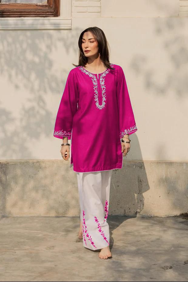 Woman wearing a bright pink embroidered top and white pants standing outdoors.