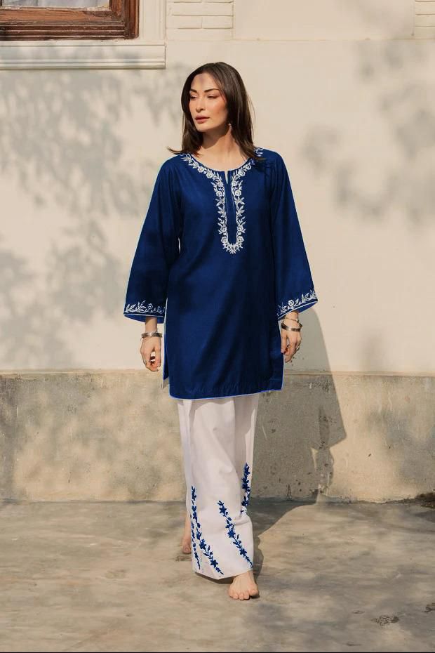 Woman wearing a blue embroidered top and white pants standing against a light-colored wall.