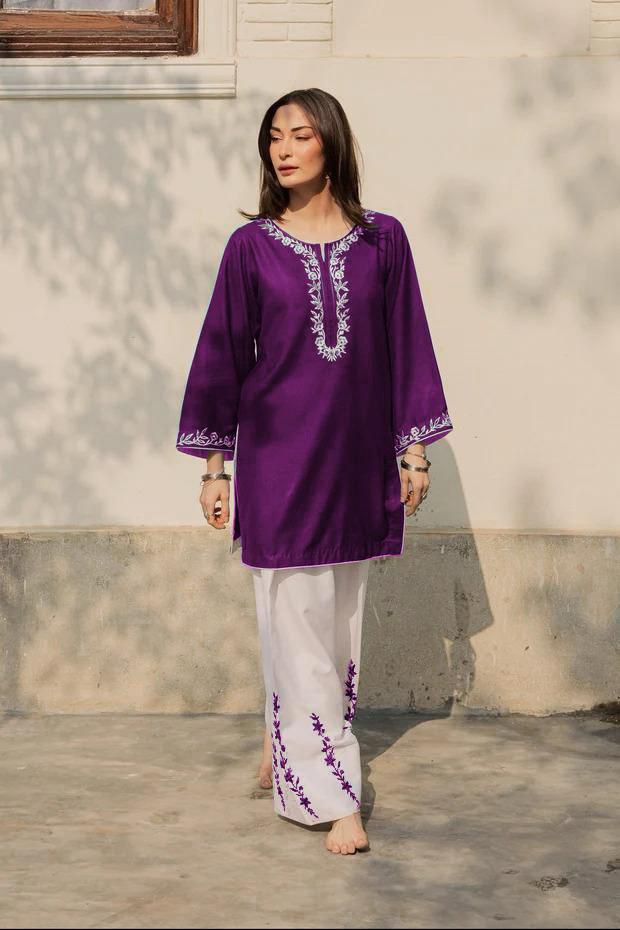 Woman wearing a purple embroidered top and white pants standing against a neutral wall.