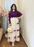 Woman in a purple sweater and white skirt with purple accents standing in a room with flowers and furniture.