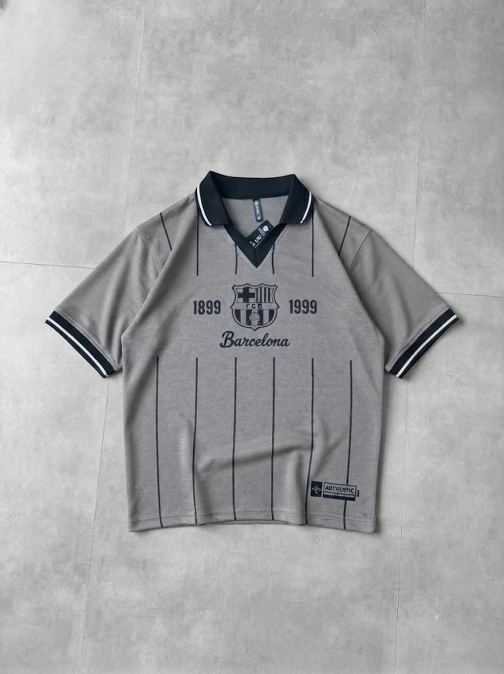 A light gray short-sleeve polo shirt with dark navy pinstripes, a dark navy collar, and 'FC Barcelona 1899 1999' printed on the chest, laid flat on a textured gray background.