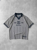 A light gray short-sleeve polo shirt with dark navy pinstripes, a dark navy collar, and 'FC Barcelona 1899 1999' printed on the chest, laid flat on a textured gray background.