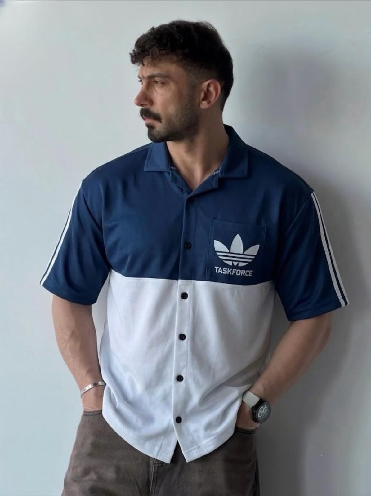 A man wearing a navy blue and white color-blocked short-sleeve button-up shirt with a white Adidas trefoil logo and 'TASKFORCE' text on the chest, standing against a plain light gray wall.