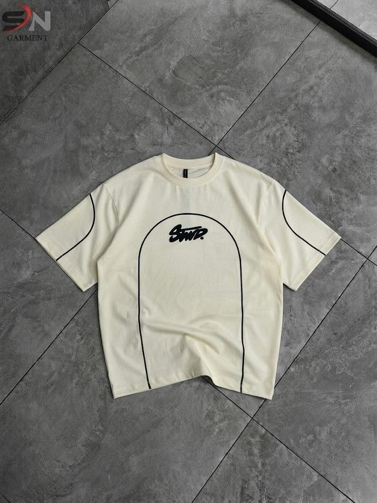 An off-white short-sleeve t-shirt with black graphic details, laid flat on a dark gray tiled floor. The shirt features a black arched graphic outline on the front with the word 'Scout' in a stylized script inside it, and thin black piping lines along the shoulders and sides.