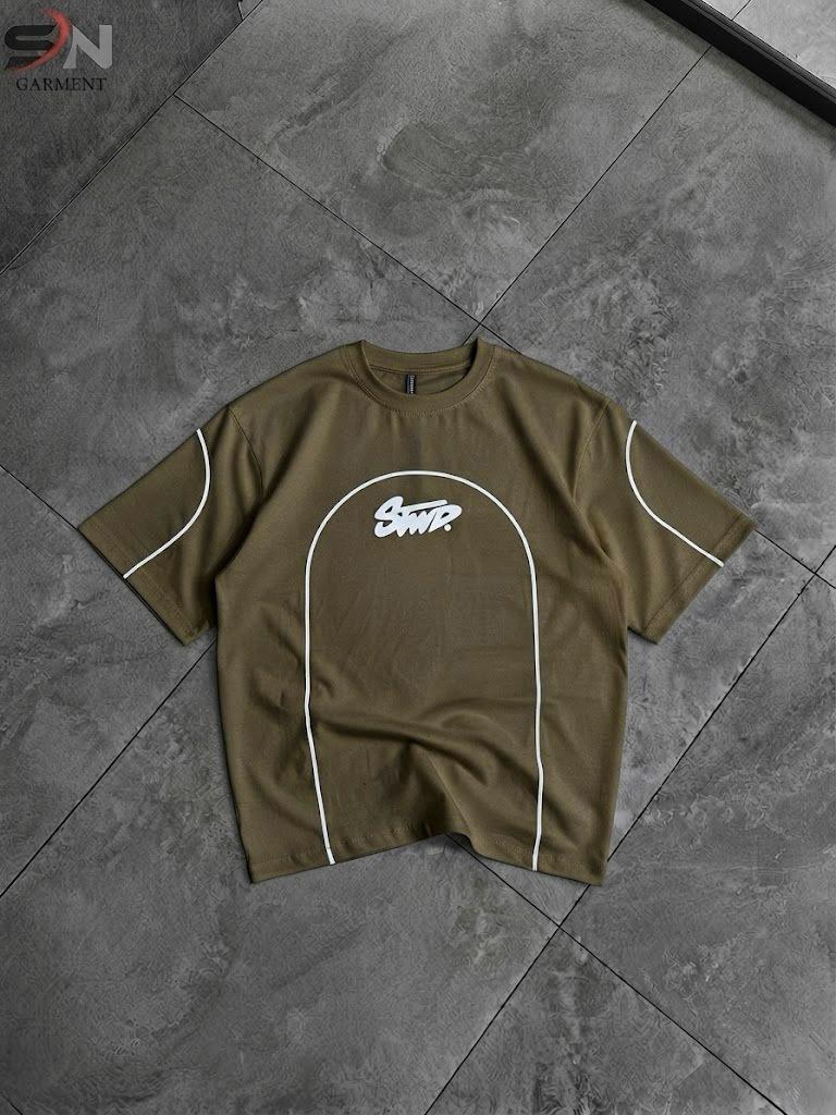 An olive green short-sleeve t-shirt with white piping and a white 'SANT' logo on the chest, laid flat on a gray tiled floor.