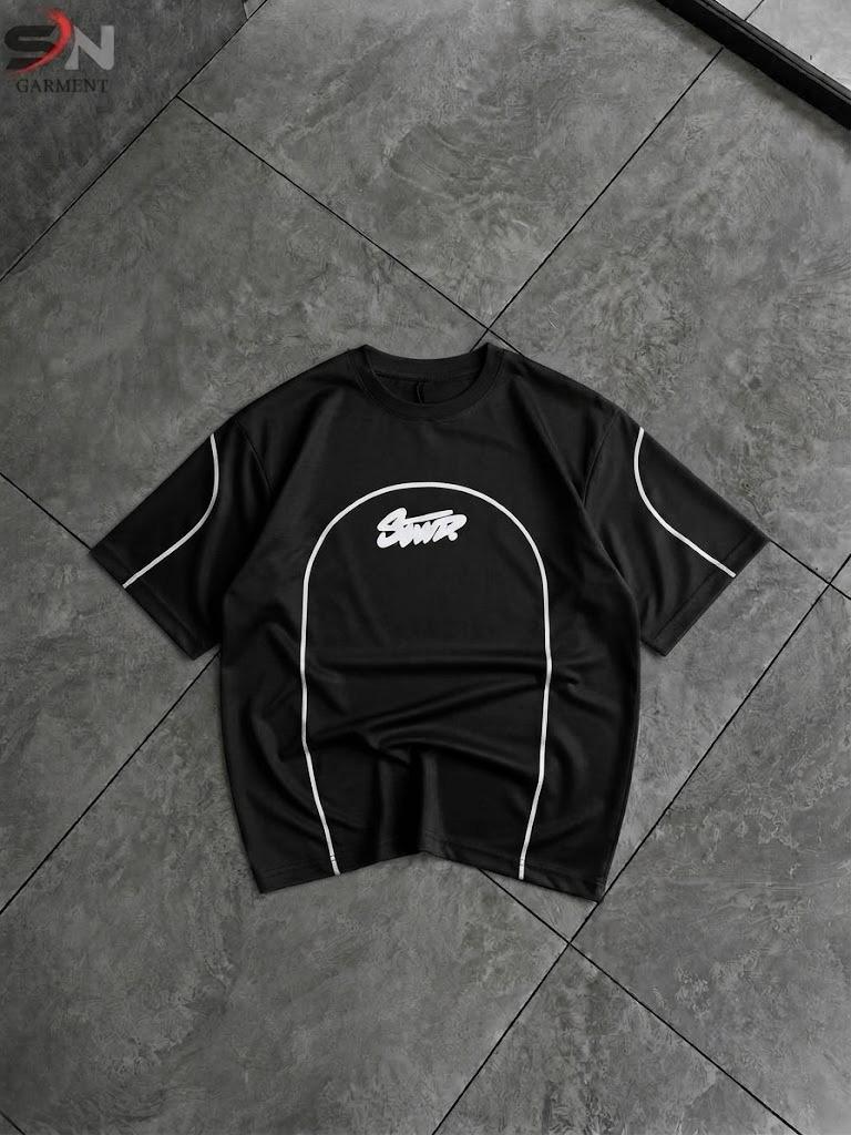 A black short-sleeve t-shirt with white contrast piping and a white graphic logo on the chest, laid flat on a dark gray tiled floor.
