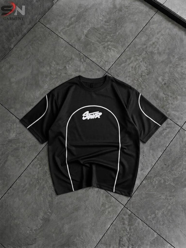 A black short-sleeve t-shirt with white contrast piping and a white graphic logo on the chest, laid flat on a dark gray tiled floor.