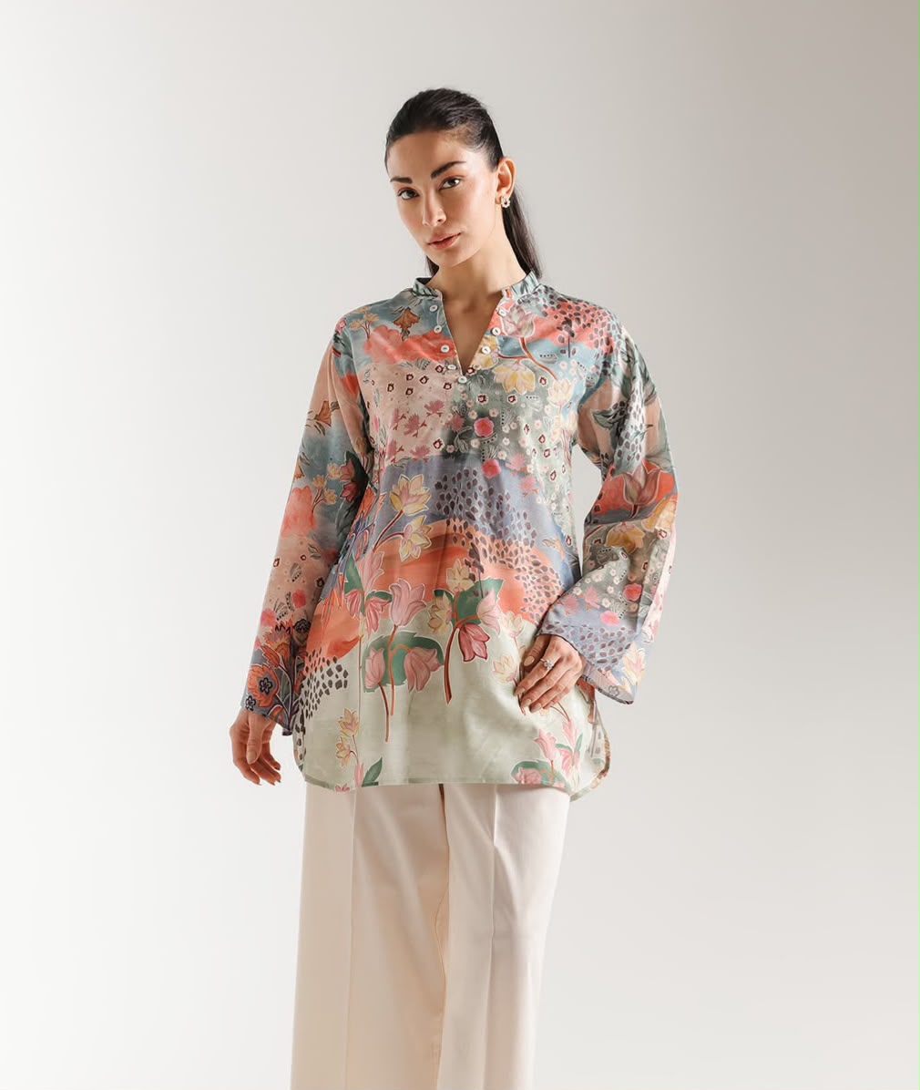 A woman modeling a long-sleeve button-up shirt with a colorful, painterly abstract pattern in coral, blue, green, and beige, paired with light beige wide-leg trousers, against a plain light gray studio background.