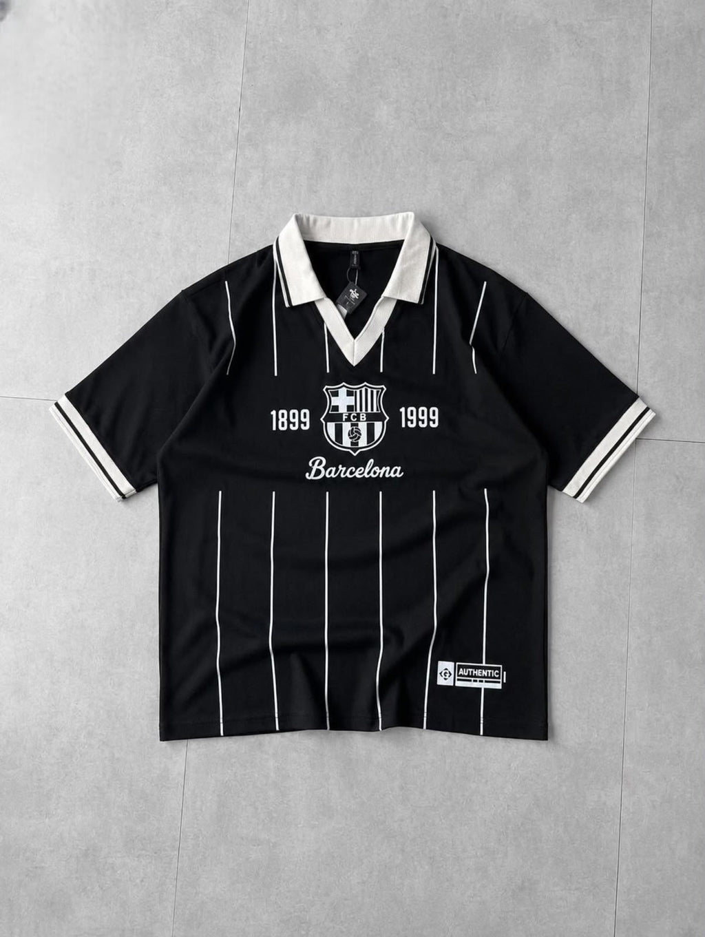 A black short-sleeve FC Barcelona soccer jersey with white pinstripes, a white collar, and white printed graphics on the chest, laid flat on a gray tiled background.