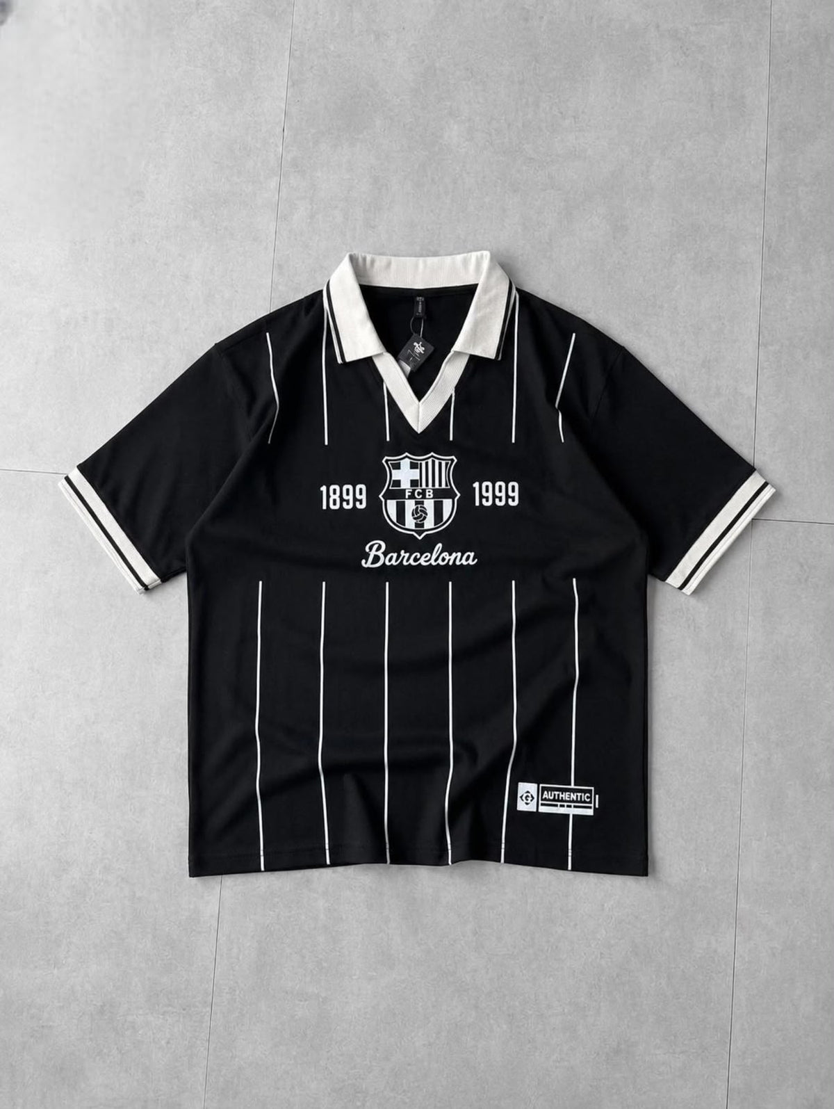 A black short-sleeve FC Barcelona soccer jersey with white pinstripes, a white collar, and white printed graphics on the chest, laid flat on a gray tiled background.