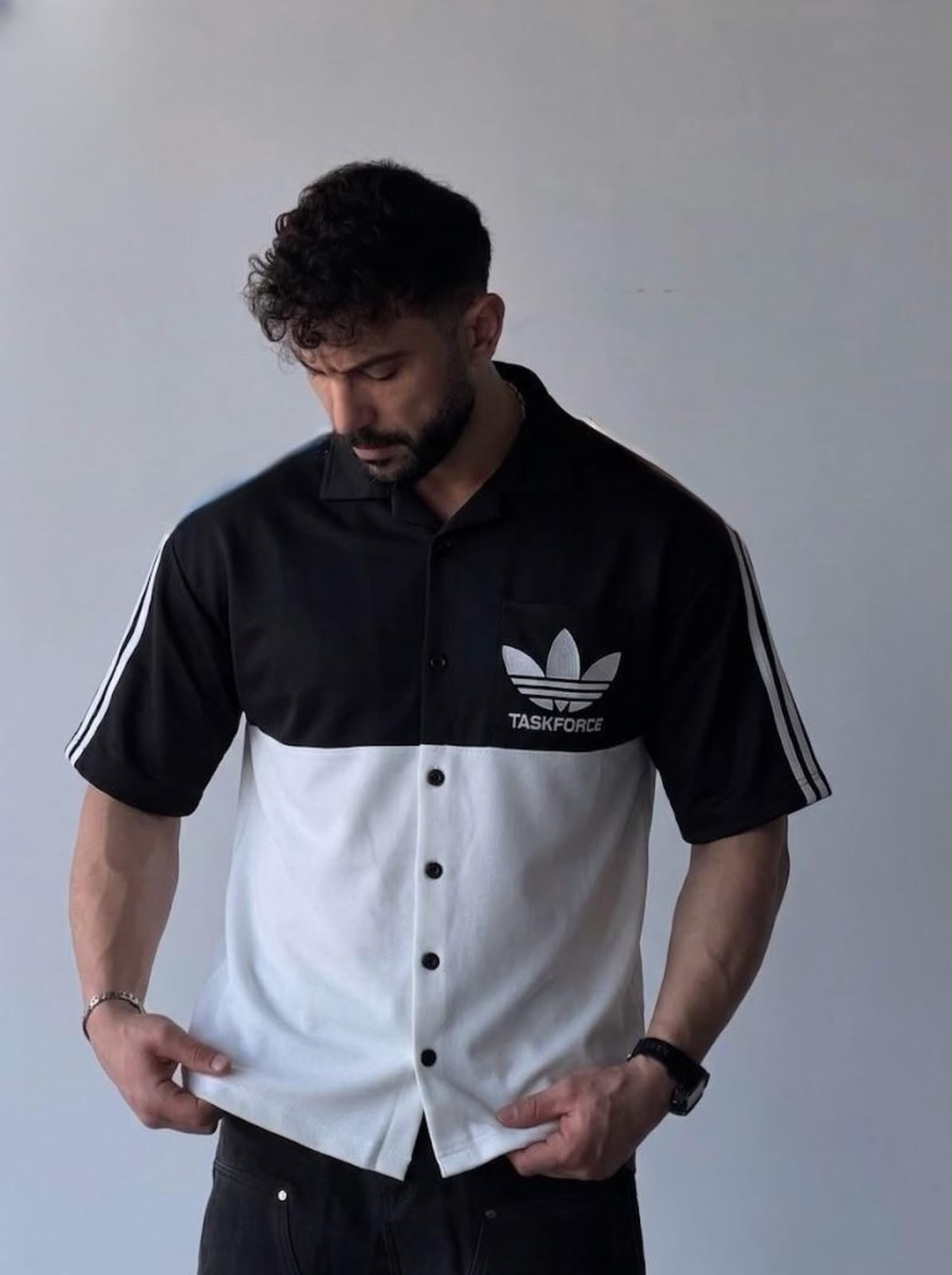 A man wearing a black and white color-blocked short-sleeve button-up shirt with the Adidas Trefoil logo and 'TAG HEURSE' text on the chest, and white stripes on the sleeves, posed against a plain gray wall.