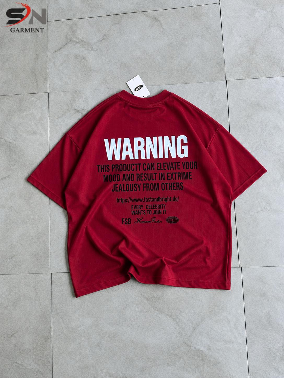 A red oversized graphic t-shirt laid flat on a light gray tiled floor, featuring a large white 'WARNING' text print on the back with smaller black text underneath.