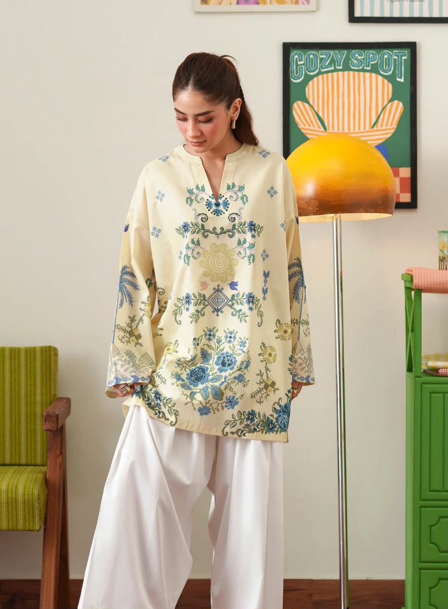 A woman wearing a cream-colored embroidered kurta with blue and green floral patterns, paired with white wide-leg trousers, standing in a decorated room with a yellow floor lamp and framed art.