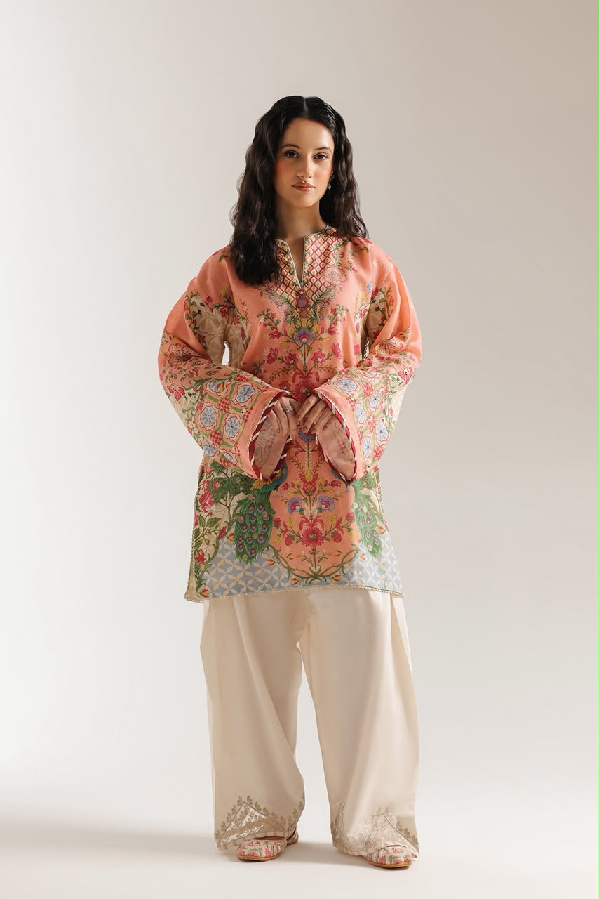 A woman modeling a traditional long-sleeve tunic top with a peach base and colorful floral embroidery, paired with cream-colored wide-leg pants, against a plain light gray studio background.