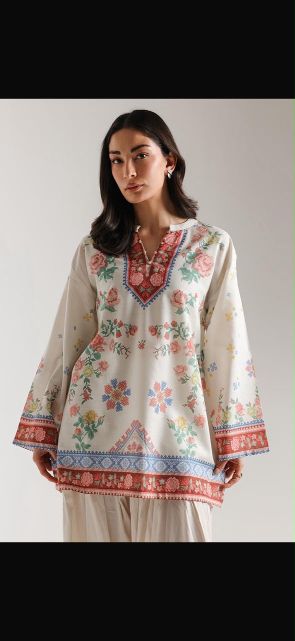A woman modeling a cream-colored kurta with colorful floral embroidery and red, blue, and green border patterns, paired with matching cream pants against a neutral studio background.