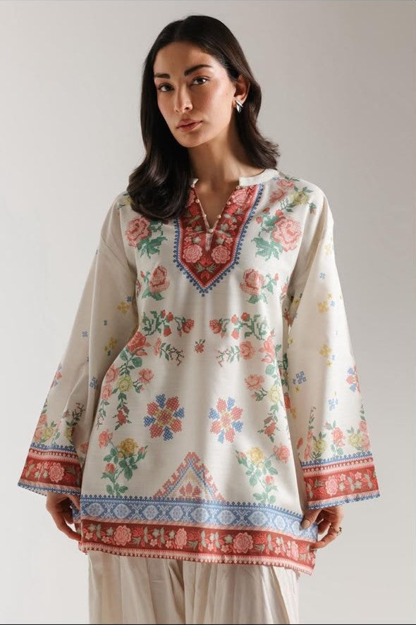 A woman modeling a cream-colored cotton tunic top with colorful floral and geometric patterns, paired with light beige trousers, against a plain light gray studio background.