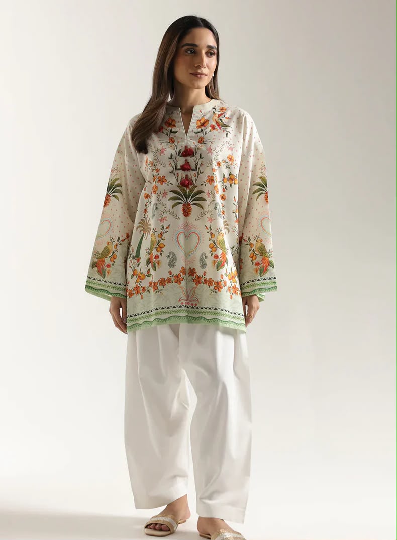 A woman modeling a cream-colored embroidered kurta with green and orange floral patterns, paired with white wide-leg trousers, against a plain light gray studio background.