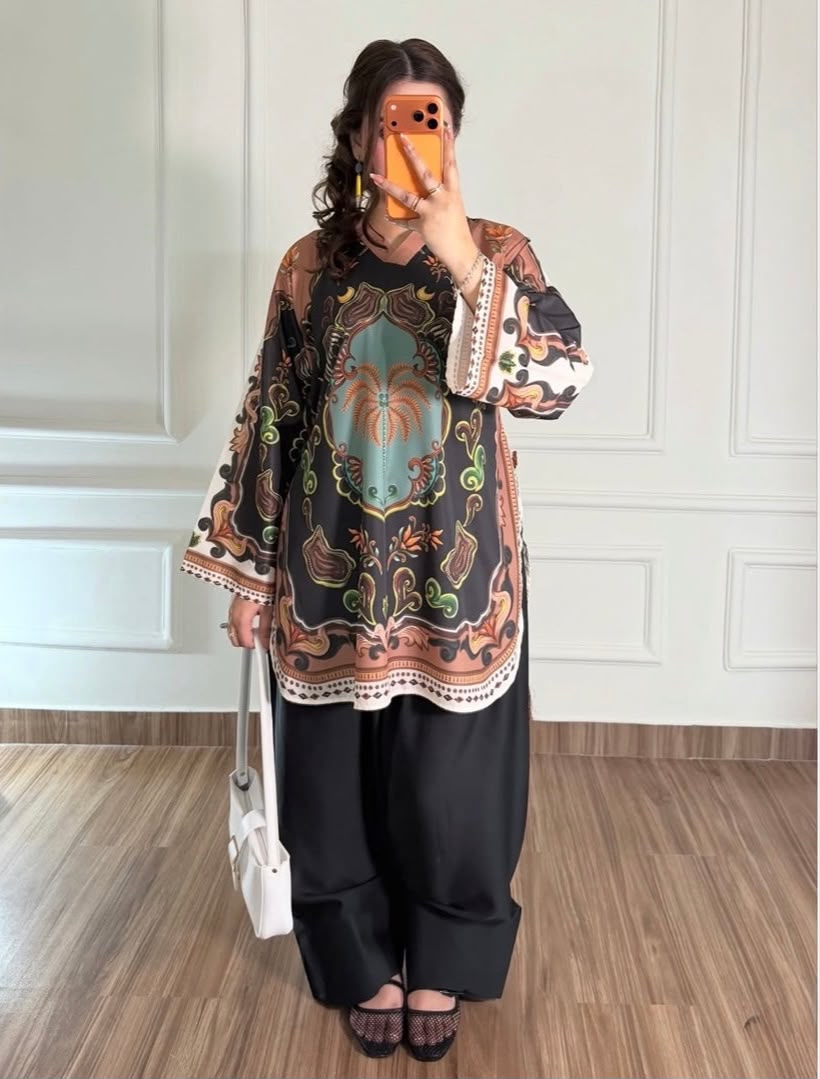 A woman takes a mirror selfie wearing a bohemian-style printed tunic top with black wide-leg pants, holding a white handbag. The tunic features a complex pattern with brown, orange, green, and teal on a black base, paired with black slip-on shoes.