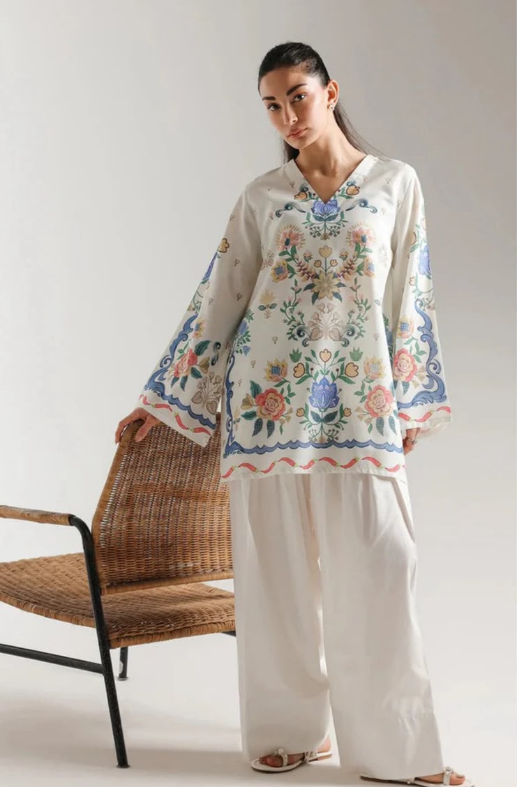 A woman models a cream-colored embroidered tunic top with wide sleeves and matching wide-leg trousers, standing next to a woven chair against a light gray studio background.