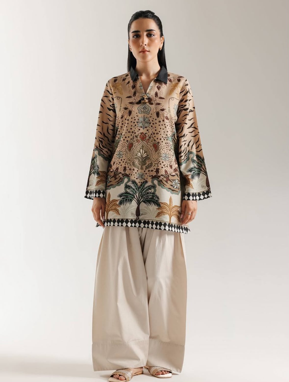 A woman modeling a traditional long-sleeve printed tunic top with wide-leg beige trousers, standing against a plain light gray studio background.