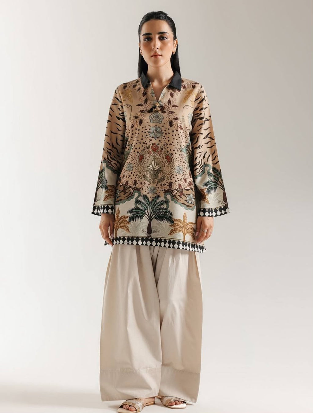 A woman modeling a traditional long-sleeve printed tunic top with wide-leg beige trousers, standing against a plain light gray studio background.